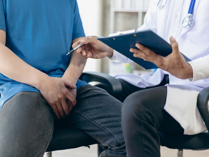 A man in a blue t shirt with black trousers is sitting on a chair to the left but you can only see his chest and upper legs. A South Asian doctor in a white medical coat holding a blue clipboard sits to his right, gesturing towards the first man,