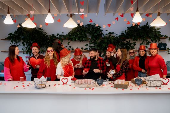 A group of male and female colleauges stood in front of a table smiling and dressed in red t shirts and sparkly hats