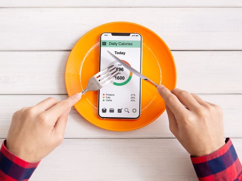 Two hands holding a knife and fork over a smartphone on an orange plate