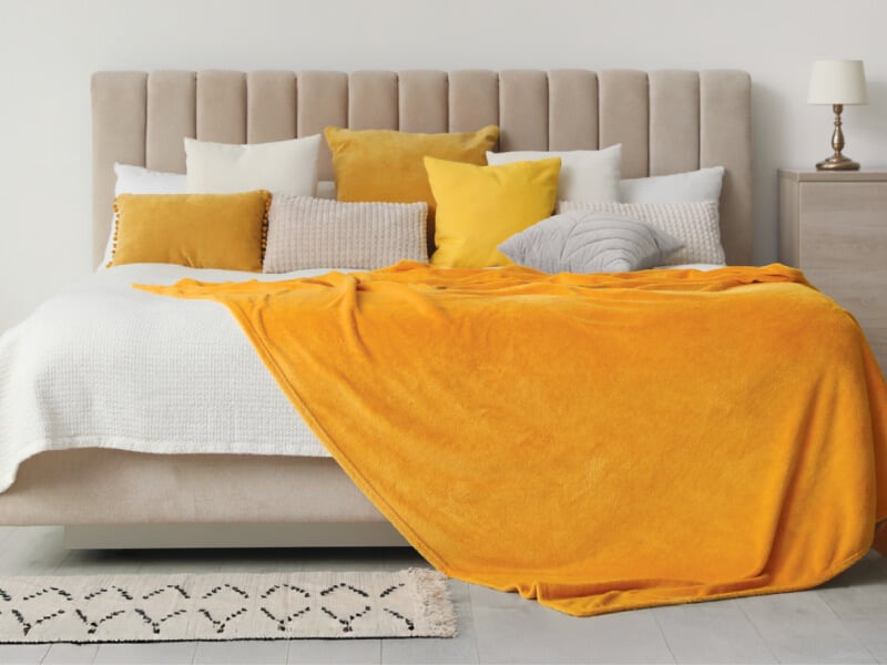 A photograph of a bed with white linen and a yellow throw with cushions.