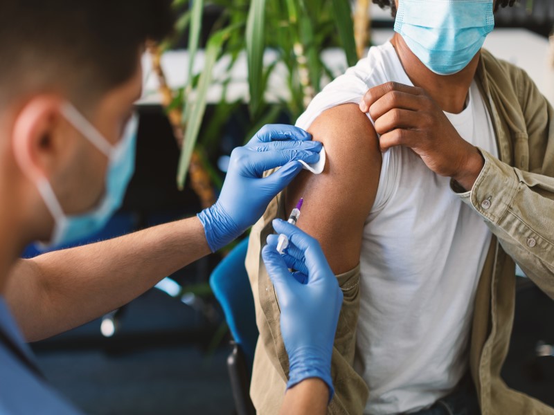 A healthcare professional in blue scrubs wearing a blue face mask and blue latex gloves holds a syringe about to give a vaccine into the right arm of a mixed race man who is also wearing a face mask