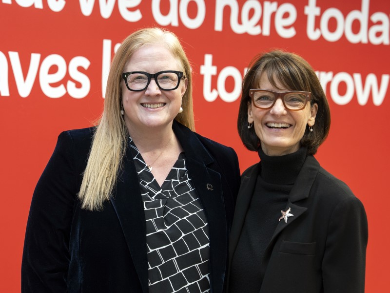 Dr Charmaine Griffiths and Dr Regina Dugan smiling in front of a red backdrop. They are both wearing black suits and black rimmed glasses,